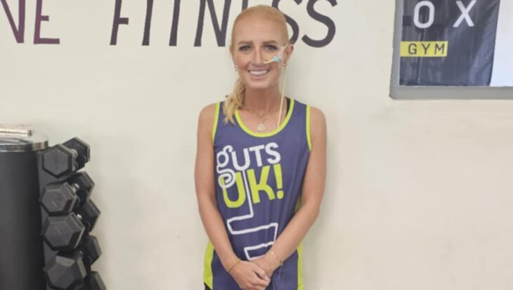 Eryn is standing in a gym wearing a blue Guts UK running vest and black shorts. She also wears black and white trainers.