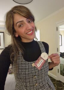 Sharan, smiling directly at the camera in a room, wearing a black turtleneck under a black and white checked pinafore dress. Attached to her dress are two badges, one of which reads "SAIA."