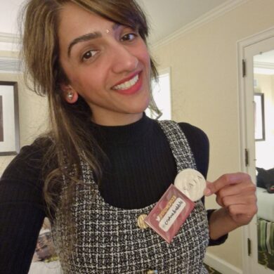 Sharan, smiling directly at the camera in a room, wearing a black turtleneck under a black and white checked pinafore dress. Attached to her dress are two badges, one of which reads