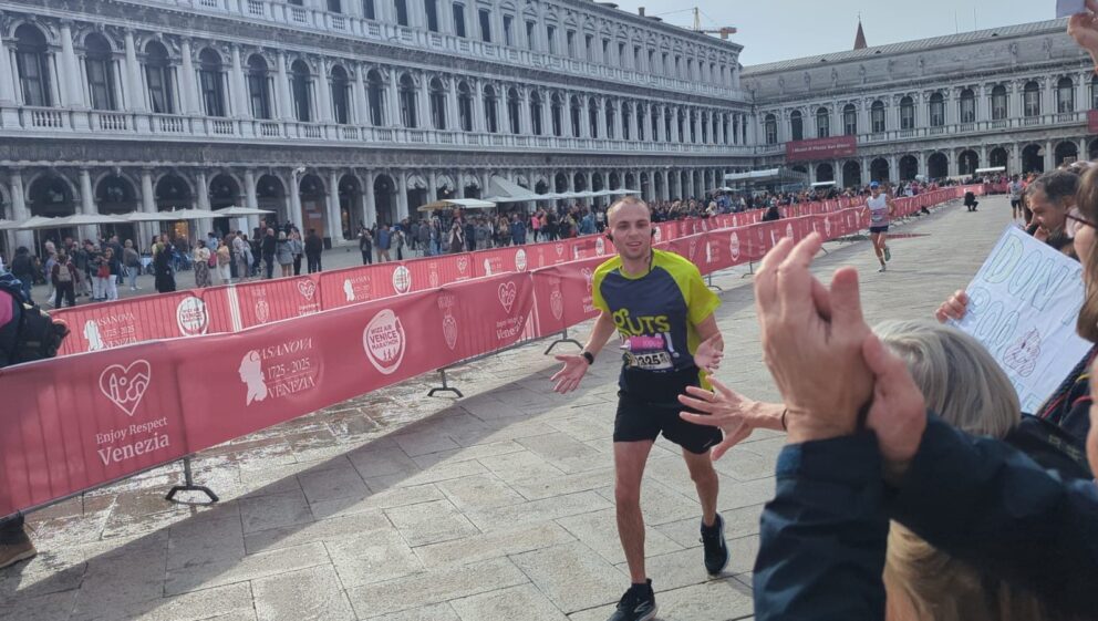 A man named Declan, wearing a Guts UK t-shirt, smiles and extends his arms whilst running the Venice Marathon.