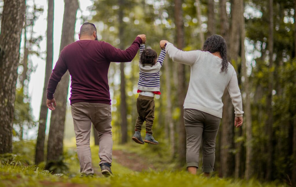  A family of three is shown from the back as they walk along a path through a wooded area. On the left, a man wears a maroon long-sleeved shirt and tan pants. On the right, a woman wears a white textured sweater and greyish-green trousers. In the center, they lift a small child into the air by their hands. The child is wearing a navy and white striped sweater, olive green pants, and grey boots. The background is filled with tall, slender trees and lush green foliage, with soft sunlight filtering through the leaves.