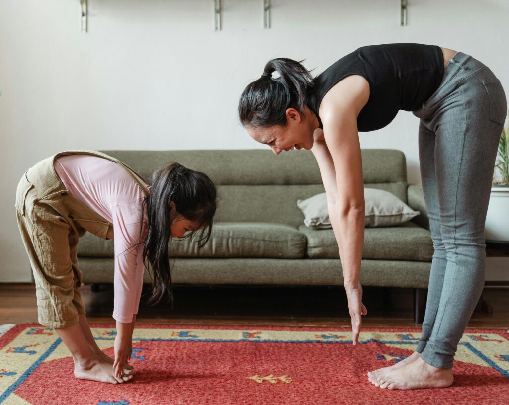  An adult and a young girl are indoors on a red patterned rug, bending forward at the waist to touch their toes. The adult, with their hair in a ponytail, wears a black sleeveless top and grey leggings. The child wears a pink long-sleeved shirt and tan overalls. They are facing each other with a green sofa in the background.