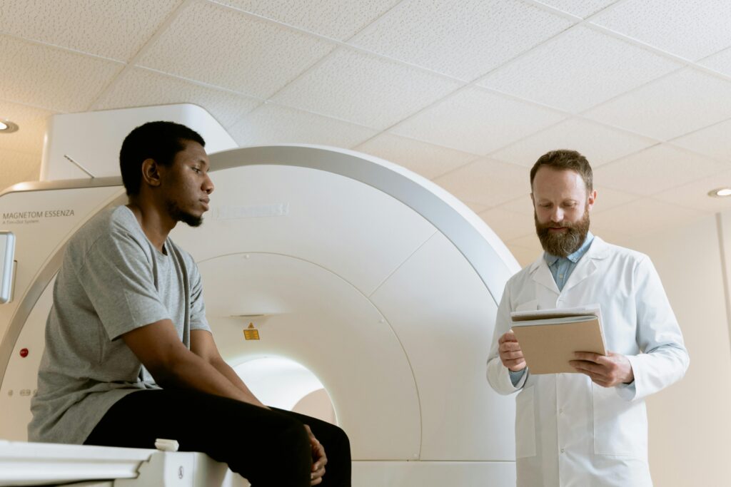 A man in a grey t-shirt and black trousers sits on the edge of a white medical scanning bed, looking toward the right. To his right, a man with a beard wearing a white lab coat stands looking down at a folder he is holding. In the background, a large, circular white MRI or CT scanner is visible under bright, indoor ceiling lights.